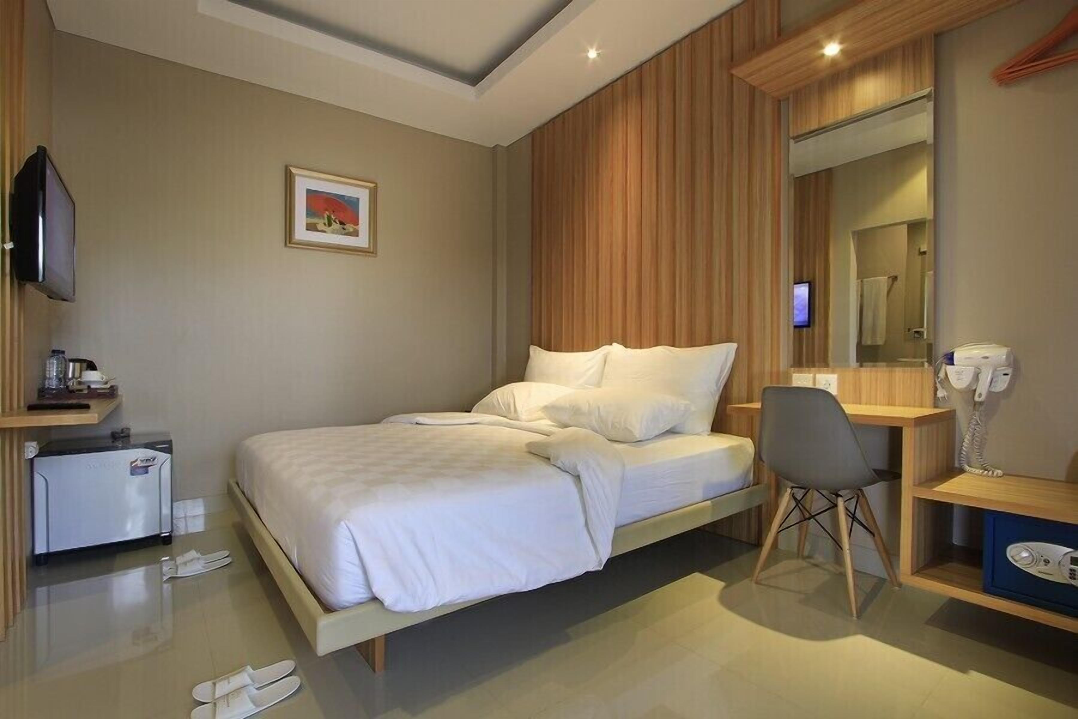 Habitación de hotel con cama, escritorio, silla, televisor, refrigerador pequeño y espejo.
