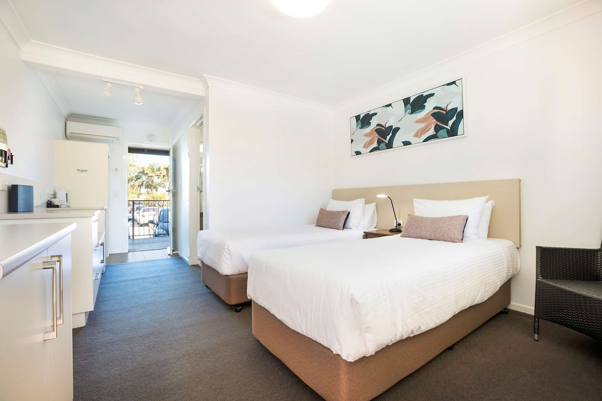 Une chambre d’hôtel avec deux lits, un bureau et une chaise. Il y a un tableau au mur et la chambre offre une vue sur l’extérieur.