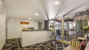 A reception area with a reception desk, a patterned carpet, a seating area with a sofa and a chair, and a glass wall offering a view of the interior. - Nightcap at Golden Beach Tavern (Caloundra)