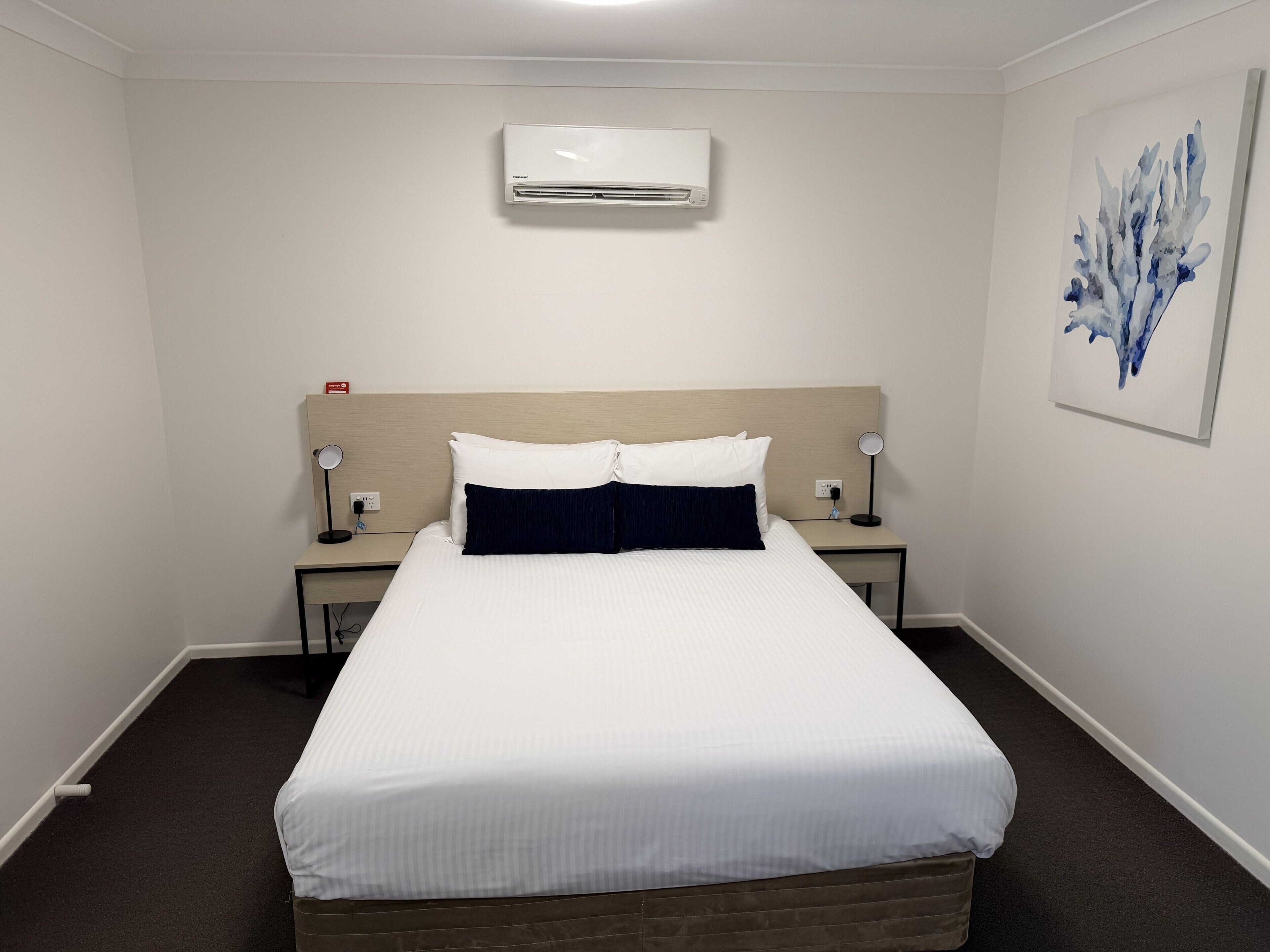 Ein Hotelzimmer mit einem Bett, zwei Nachttischen mit Lampen, einer Klimaanlage und einem Wandbild.