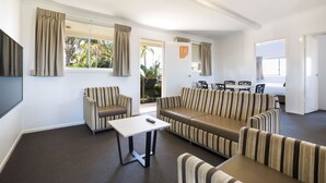 A modern living room with striped sofas, a small table, and a TV. There is a door with a number sign, and a window with curtains. - Nightcap at Golden Beach Tavern (Caloundra)