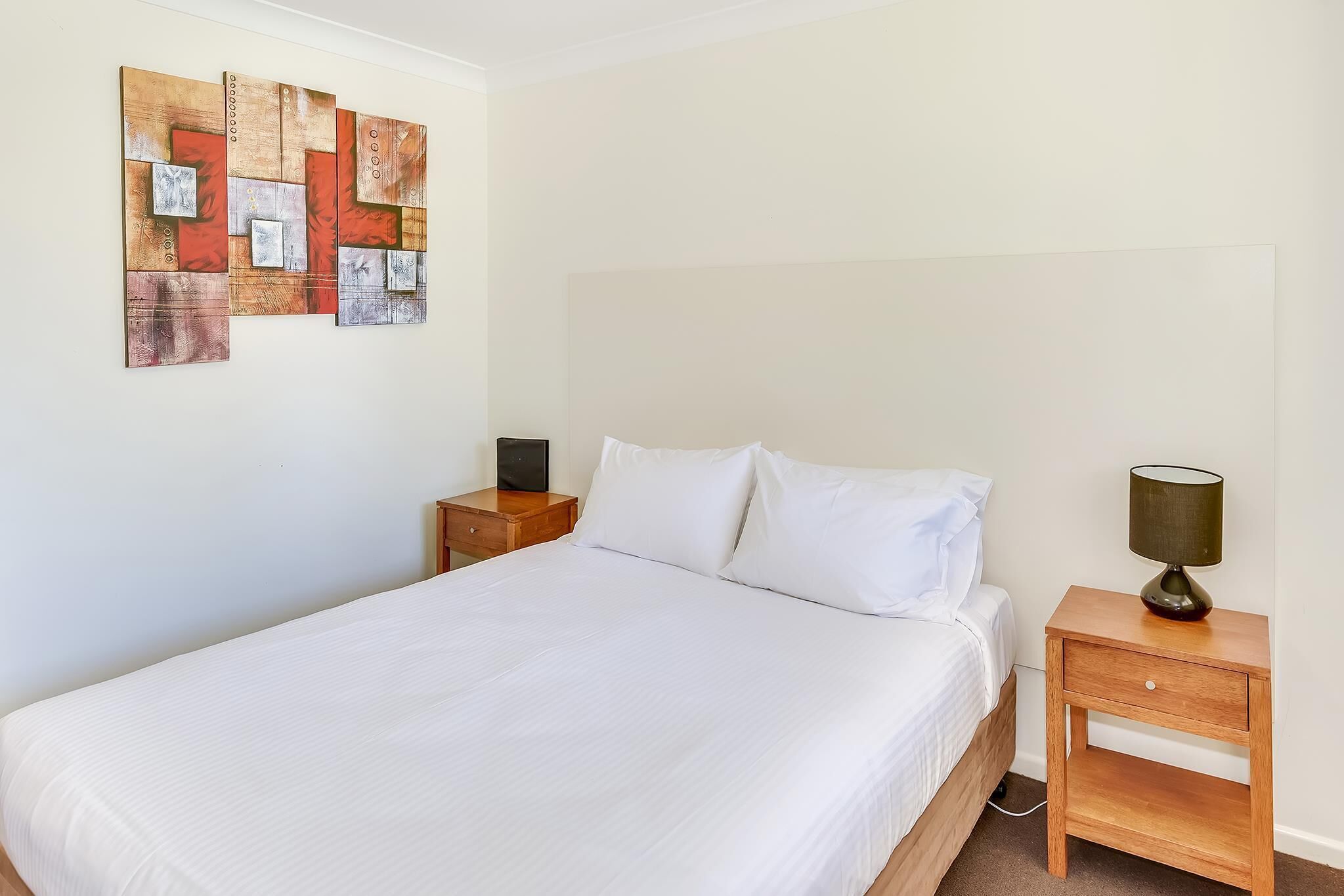 Una cama bien tendida con sábanas blancas, una mesita de noche de madera con lámpara, y una obra de arte en la pared sobre la cama.