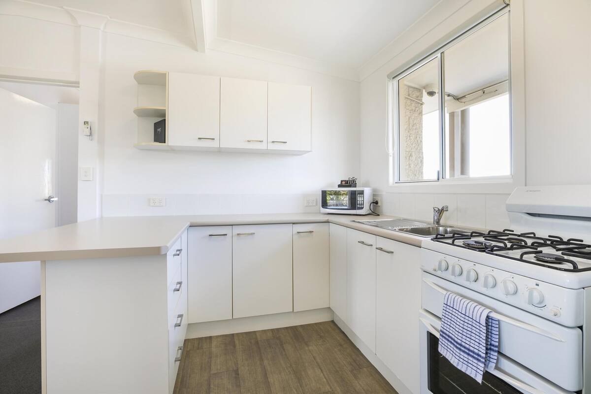 Une cuisine de taille réduite avec des meubles blancs, un four à micro-ondes, une cuisinière et une fenêtre.