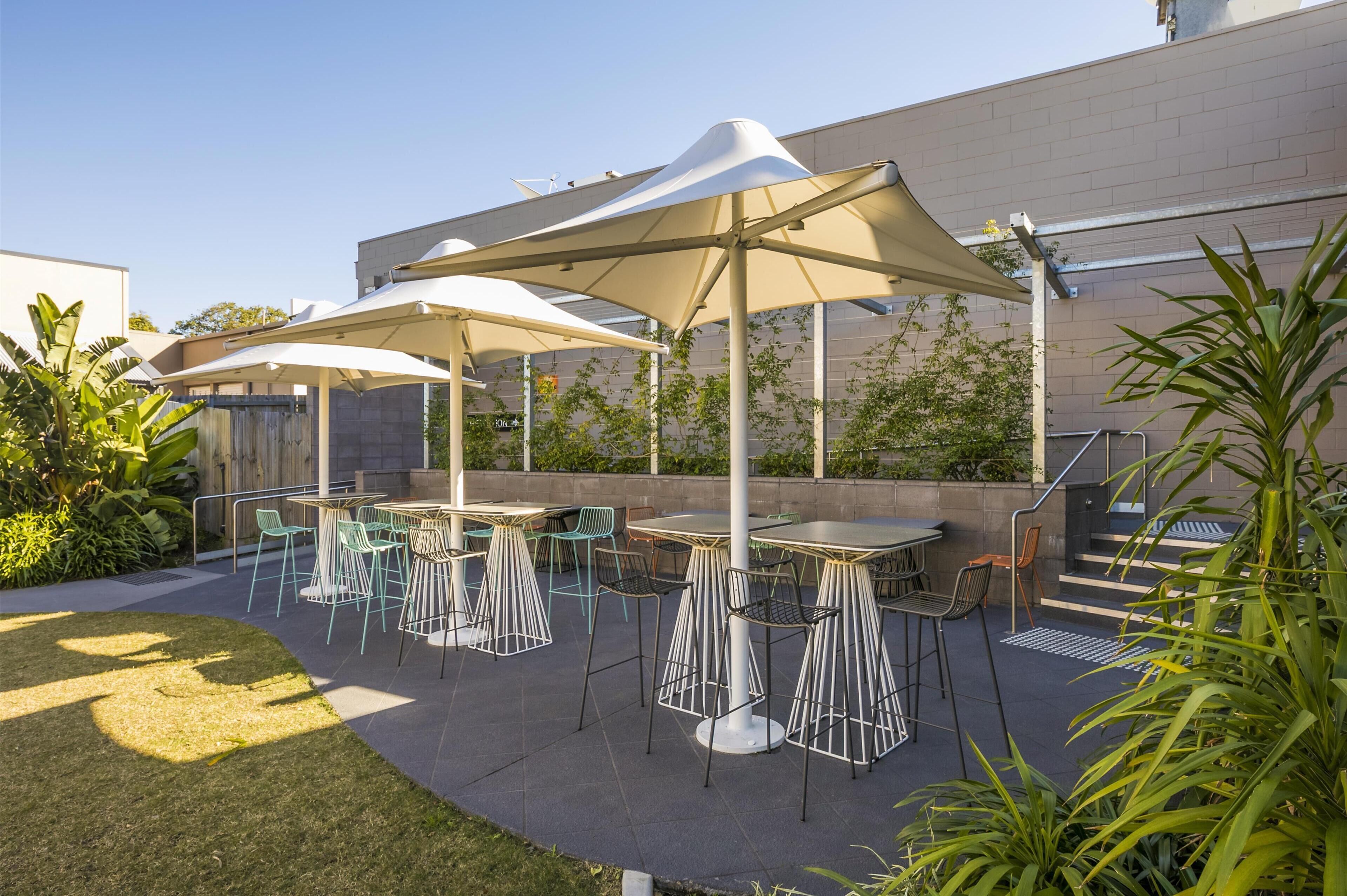 Eine Terrassengaststätte mit Tischen und Stühlen unter Sonnenschirmen, umgeben von Pflanzen und einer Gebäudefassade.
