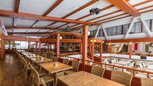 Een ruime restaurantruimte met houten tafels en rieten stoelen, hoge plafonds met zichtbare balken en grote ramen.