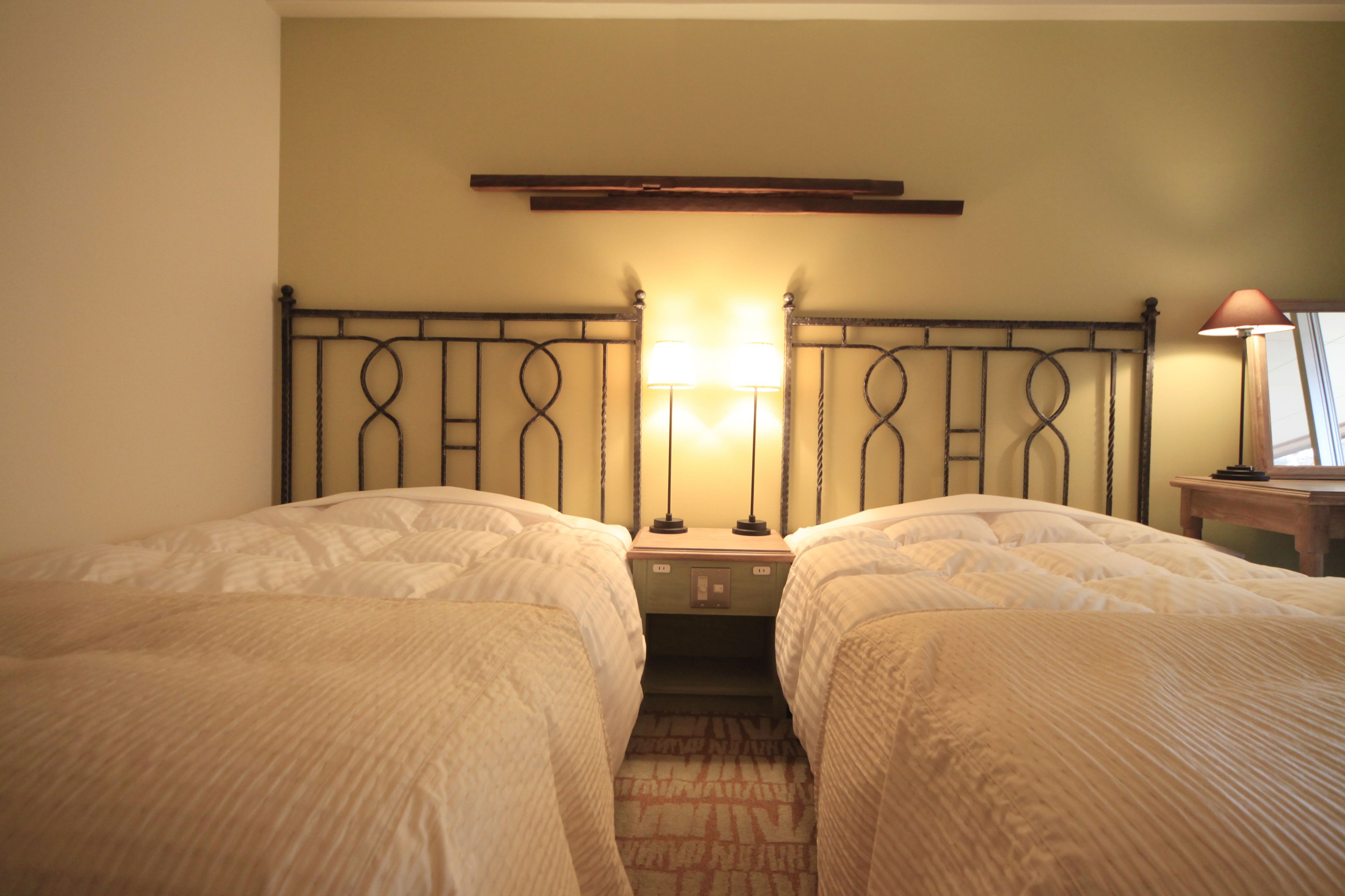 A hotel room with two single beds, a nightstand with a lamp, a wooden headboard, and a small table with a lamp.