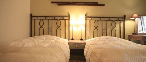A hotel room with two single beds, a nightstand with a lamp, a wooden headboard, and a small table with a lamp.