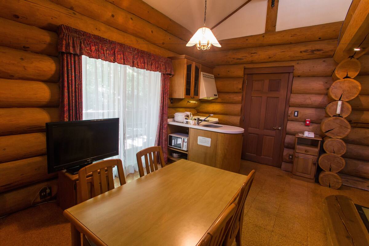 Interieur van een blokhut met een houten eettafel, stoelen, een televisie, een keuken met een magnetron en een houten kast.