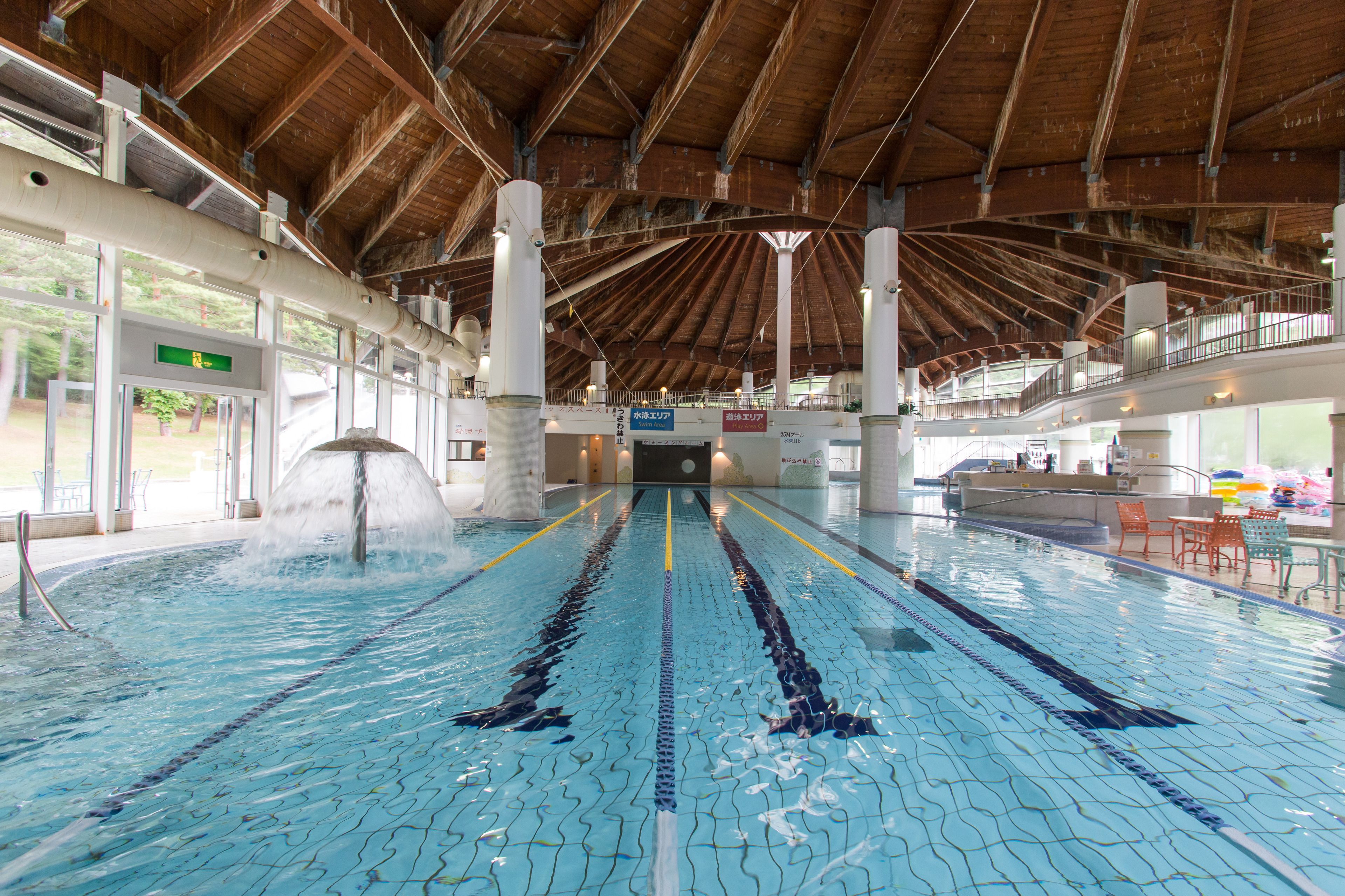 An indoor swimming pool with clear lanes, a wooden ceiling, and a reception area with chairs and tables.