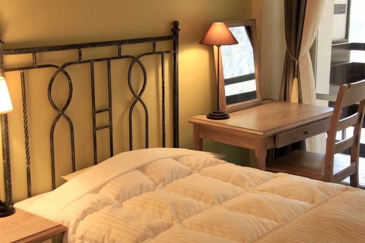 A bed with a quilted white duvet, a wooden nightstand with a lamp, a wooden desk with a mirror, and a chair.