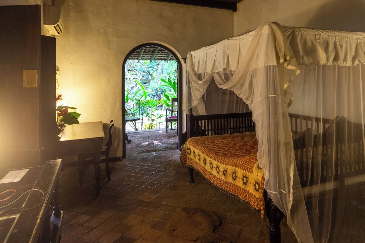 A room with a bed under a canopy, a ceiling fan, and an arched doorway leading outside.