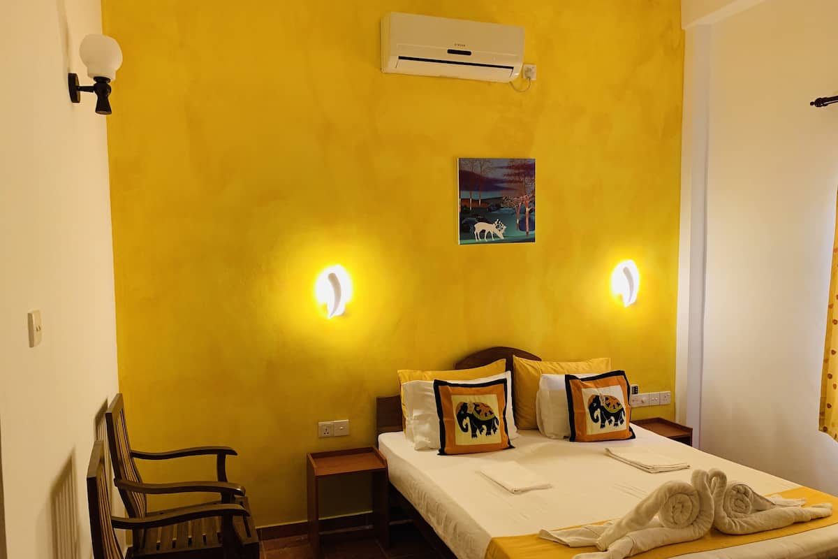 Une chambre d’hôtel avec un mur jaune, un lit avec une literie blanche et des oreillers à motifs animaliers, une table de chevet, une chaise et un appareil de climatisation.