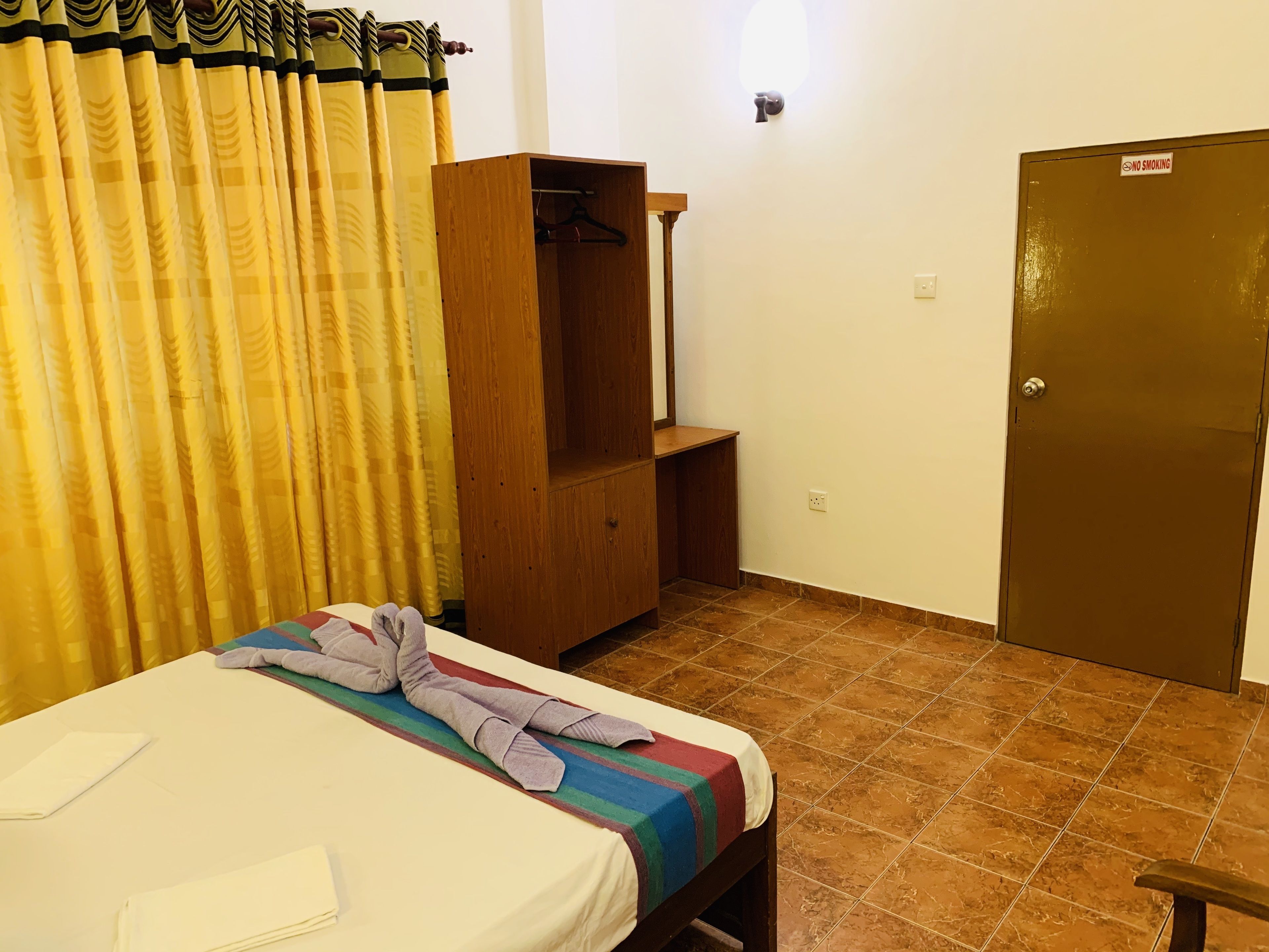 A single bed with a striped blanket, a wooden wardrobe, a coat rack, and a door with a 'No Smoking' sign.