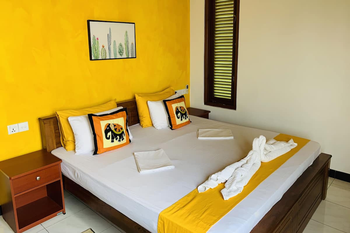Une chambre d’hôtel comprenant un lit, des oreillers, une table de chevet, une fenêtre avec des stores et une œuvre d’art accrochée au mur.