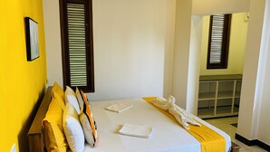 A neatly made bed with white and yellow linens, a wooden headboard, and a yellow accent pillow. There are two windows with blinds, one on each side of the bed. - Sam's Guest House (Unawatuna)