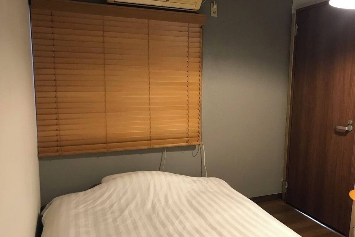 A bedroom with a bed, a window with blinds, a wall-mounted air conditioner, and a wooden door.