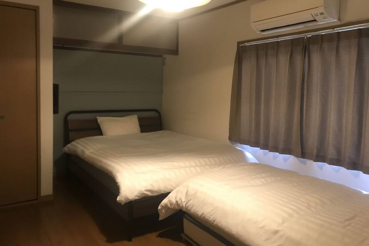 A small, single-bed room with a wooden headboard, a window with curtains, and an air conditioning unit mounted on the wall.