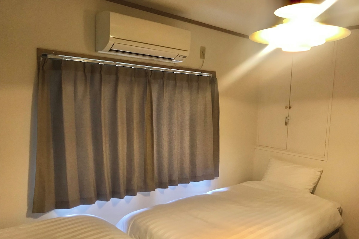 A room with two beds, an air conditioning unit, a window with curtains, and a ceiling light.