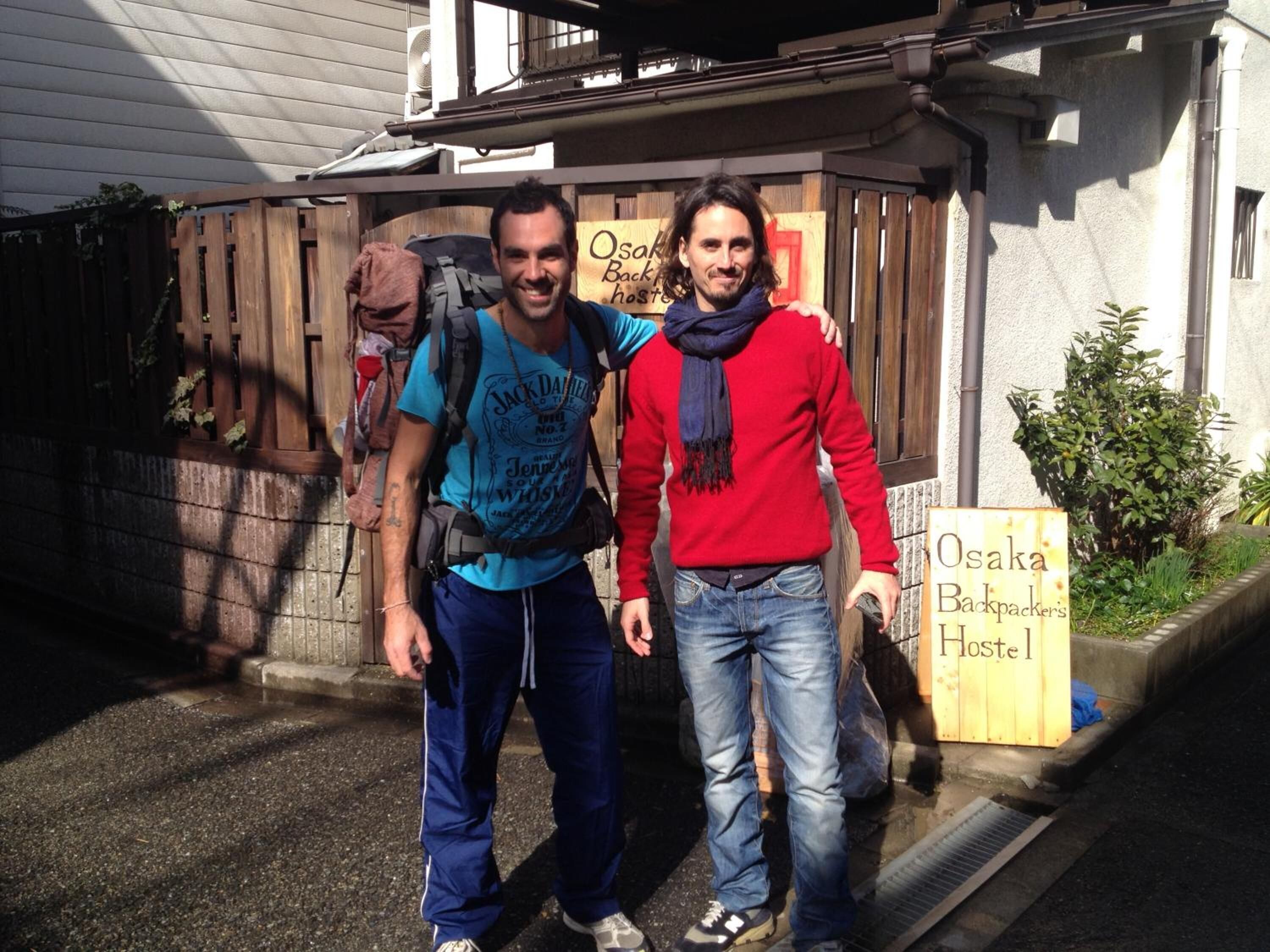 Two people standing in front of a wooden structure with a sign that reads "Osaka Backpackers Hostel".