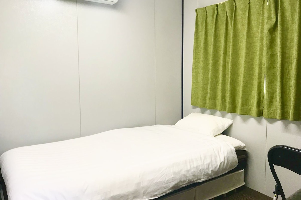 A single bed with white bedding in a room with white walls and a green curtain.