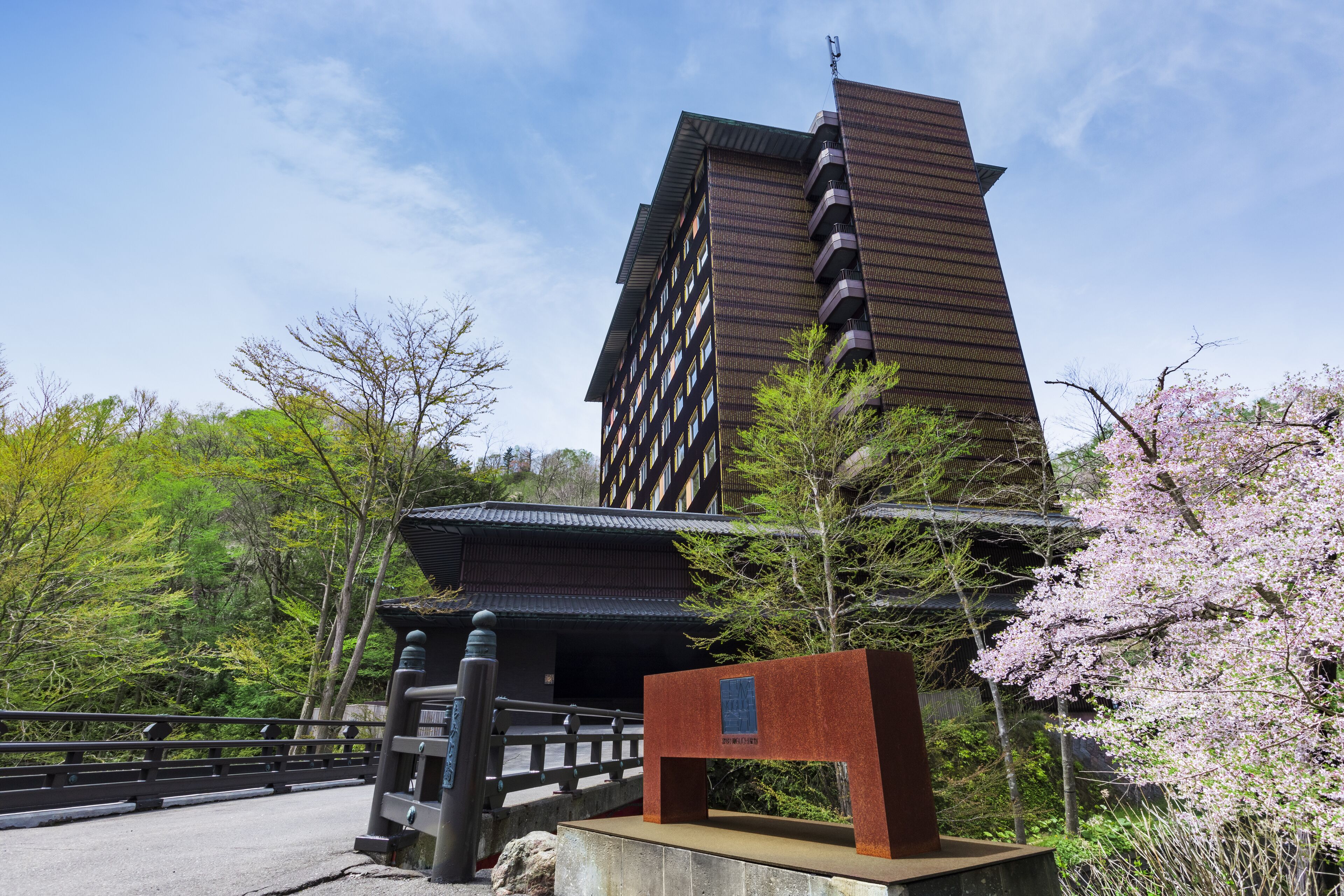 Um edifício japonês tradicional com uma extensão moderna, rodeado por vegetação exuberante e cerejeiras em flor.