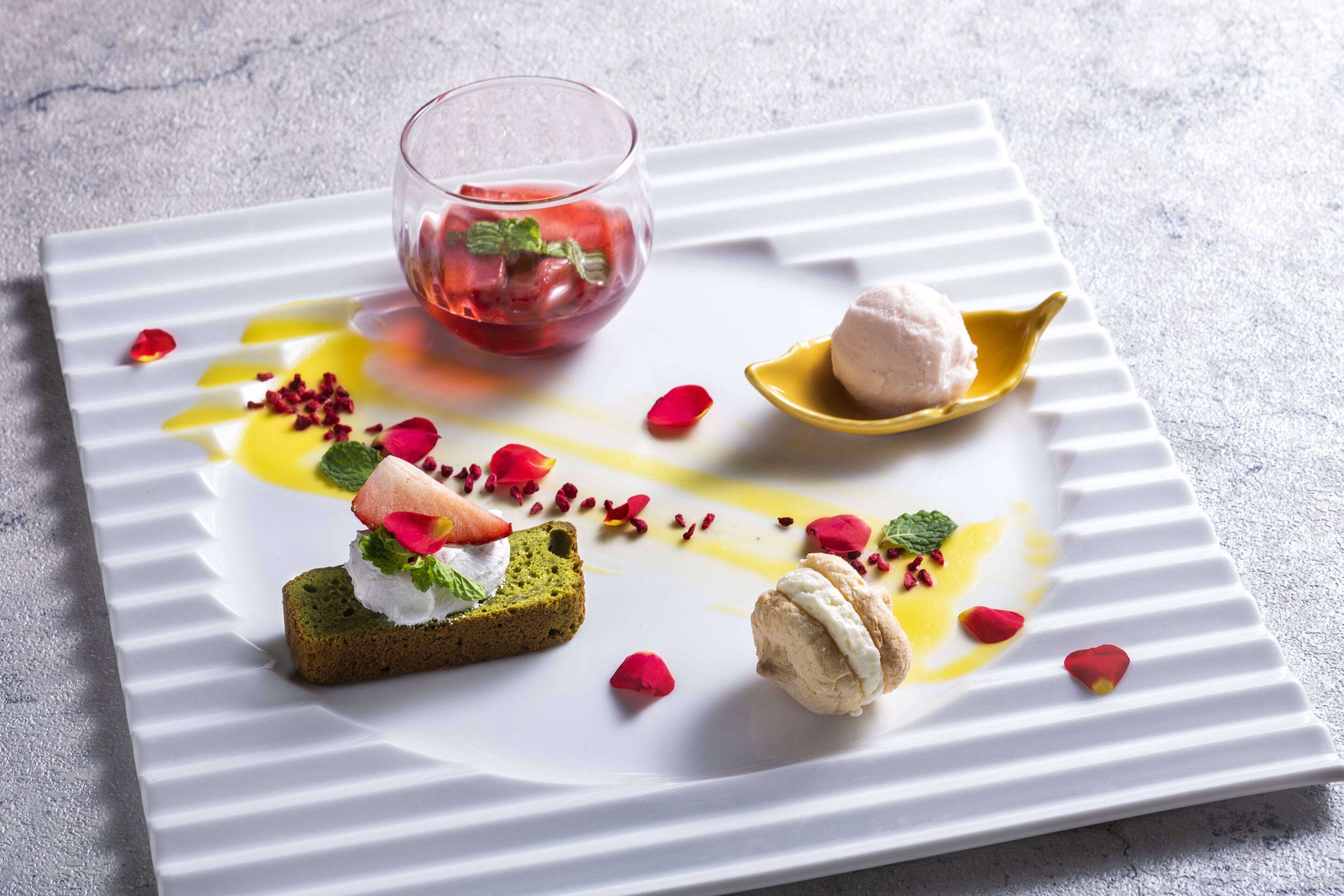 A square white plate with a dessert arrangement including a green cake, a scoop of ice cream, and a glass of red liquid.