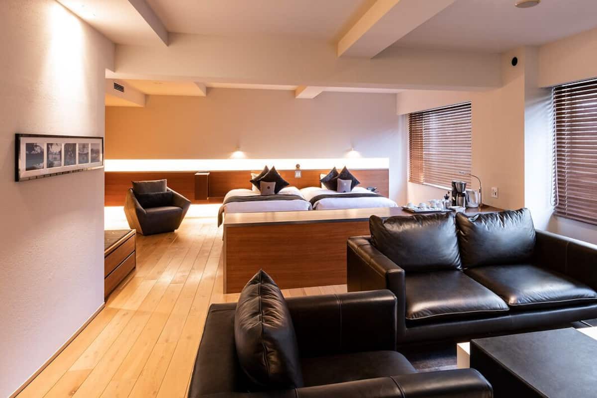 A modern hotel room with a wooden floor, a sofa, a bed with pillows, a small table, and a window with blinds.