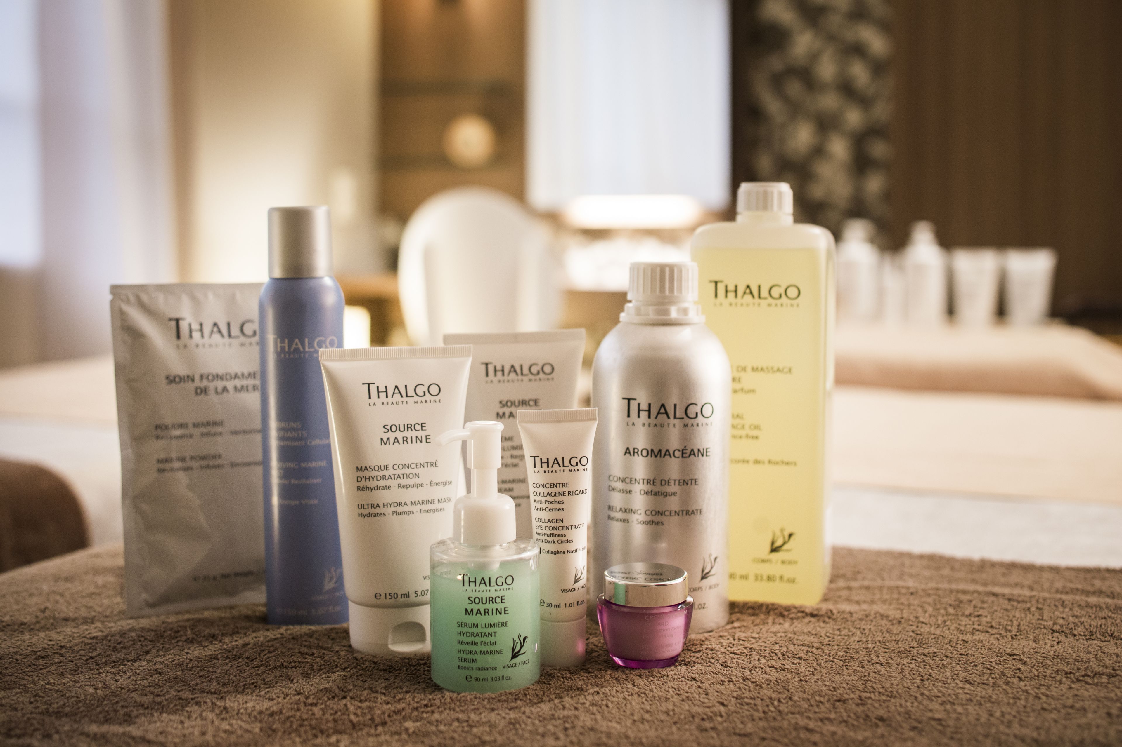 A variety of Thalgo skincare products arranged on a carpeted surface in a room with a bed and a window in the background.