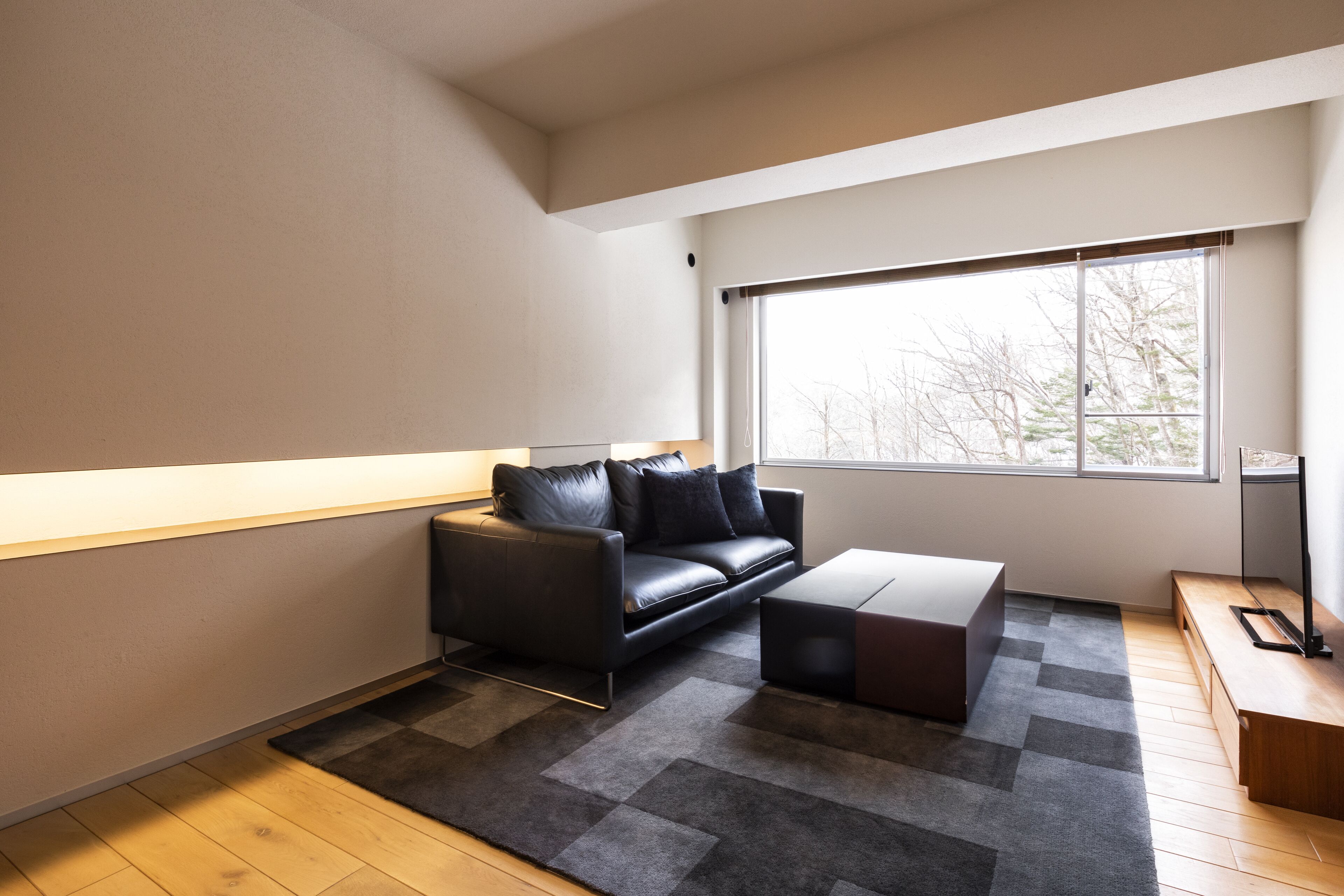 A modern living room with a black leather sofa, a wooden coffee table, and a large window providing a view of trees outside.