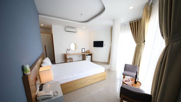 A hotel room with a large bed, a desk with a chair, a TV mounted on the wall, and a window with curtains. - Sailing Hotel (Phu Quoc)