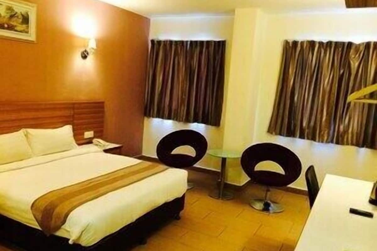 A hotel room with a bed, a desk, two chairs, a painting on the wall, and curtains on the windows.