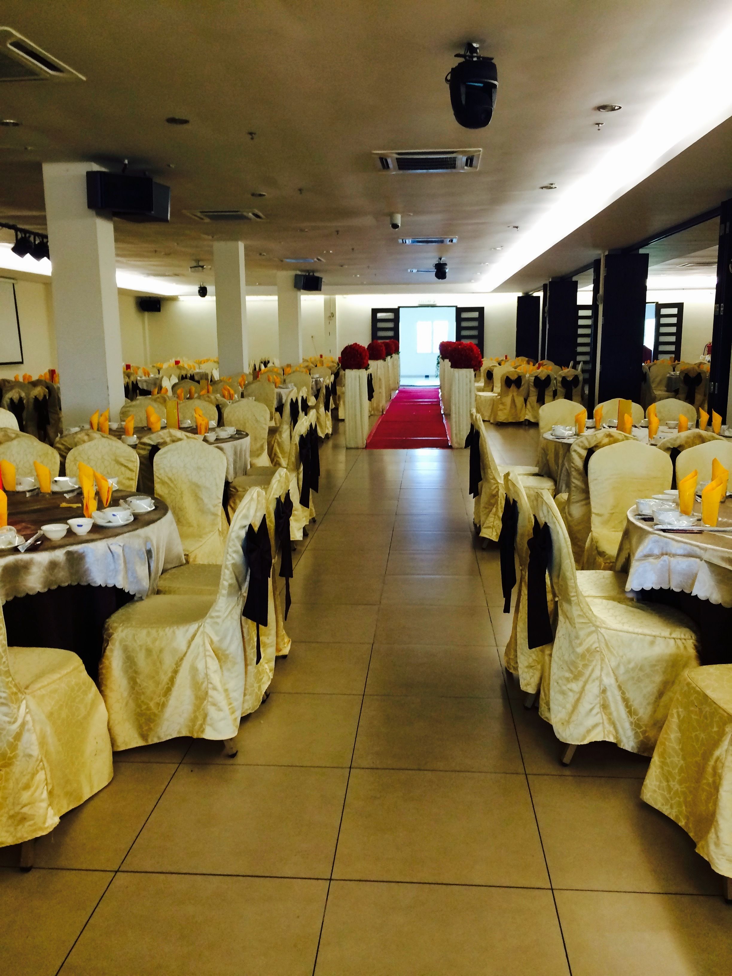 Un salón de banquetes con filas de mesas cubiertas con manteles blancos y sillas adornadas con cintas amarillas. Una alfombra roja recorre el pasillo central.