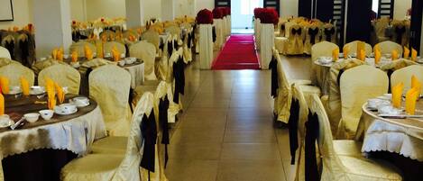 Un salón de banquetes con filas de mesas cubiertas con manteles blancos y sillas adornadas con cintas amarillas. Una alfombra roja recorre el pasillo central.