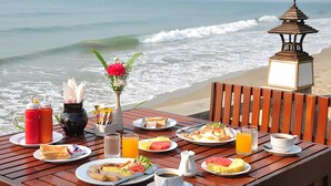 Una mesa de madera dispuesta con alimentos para el desayuno, un jarrón con flores y vistas al océano y a la playa.
