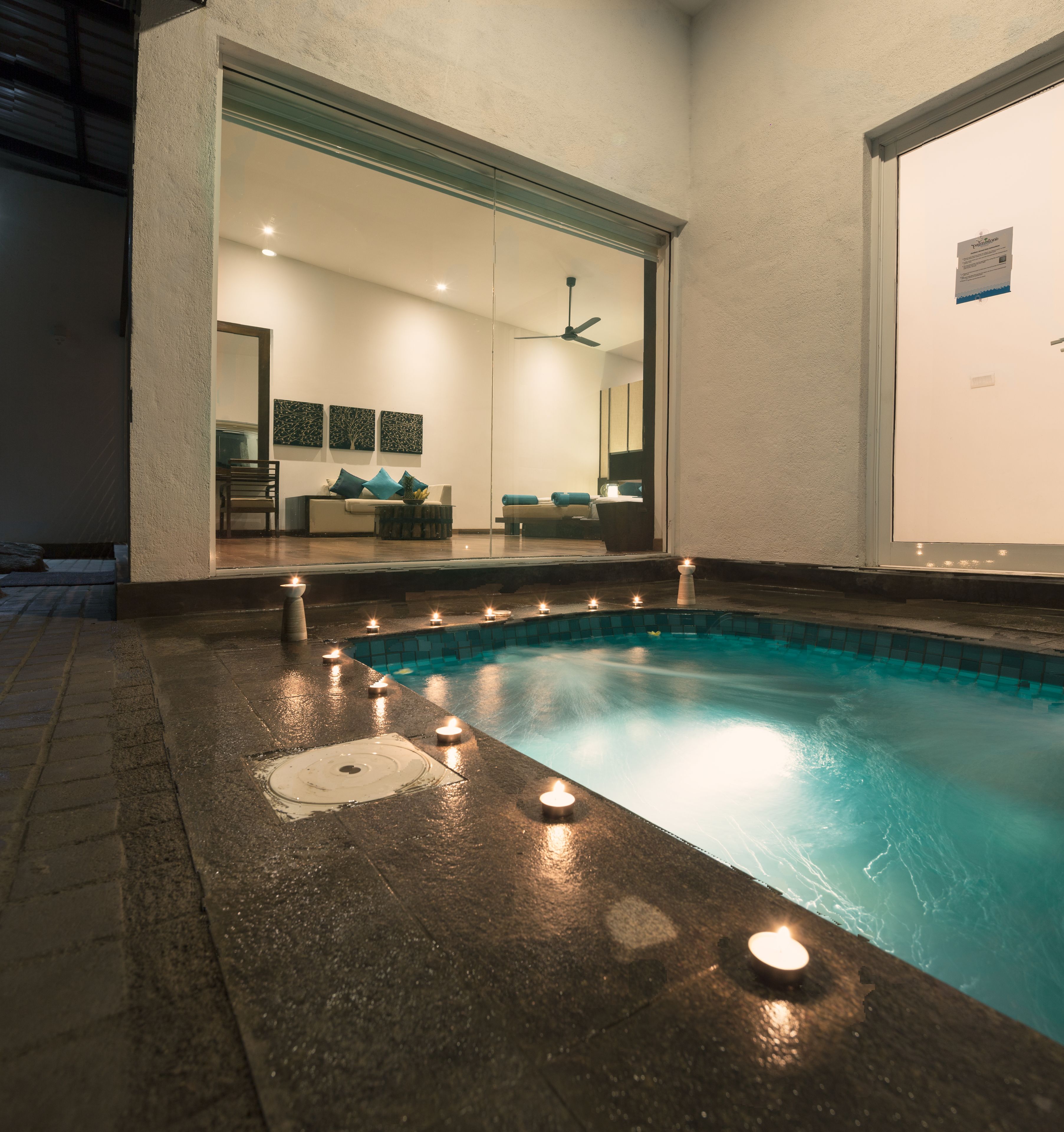 Una piscina amb espelmes al voltant del bord, una zona d'estar amb sofà i cadires, i una porta de vidre que dona a una habitació amb ventilador de sostre.