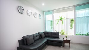 A modern living room with a black sectional sofa, a small wooden side table, and a large window with a view of greenery outside. - Su 22 (Bangkok)