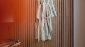 A room with a wooden slat wall, a green and white striped bathrobe hanging on a hook, and a small shelf with books. - weila - dein Boutiquehotel (Arnsberg)