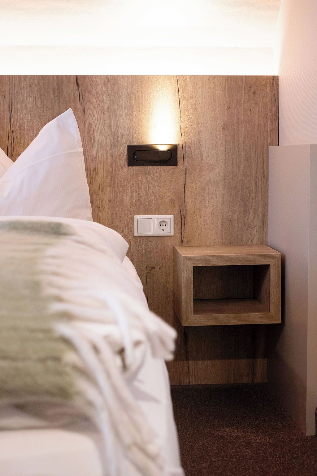 Una cama bien hecha con ropa de cama blanca, una mesita de noche con toma de corriente e iluminación de pared.