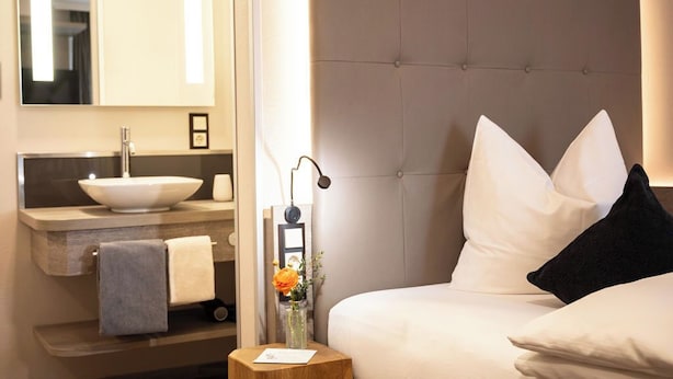 A hotel room with a large bed, a headboard, a bedside table with a lamp, a mirror, a sink, and a towel. - weila - dein Boutiquehotel (Arnsberg)