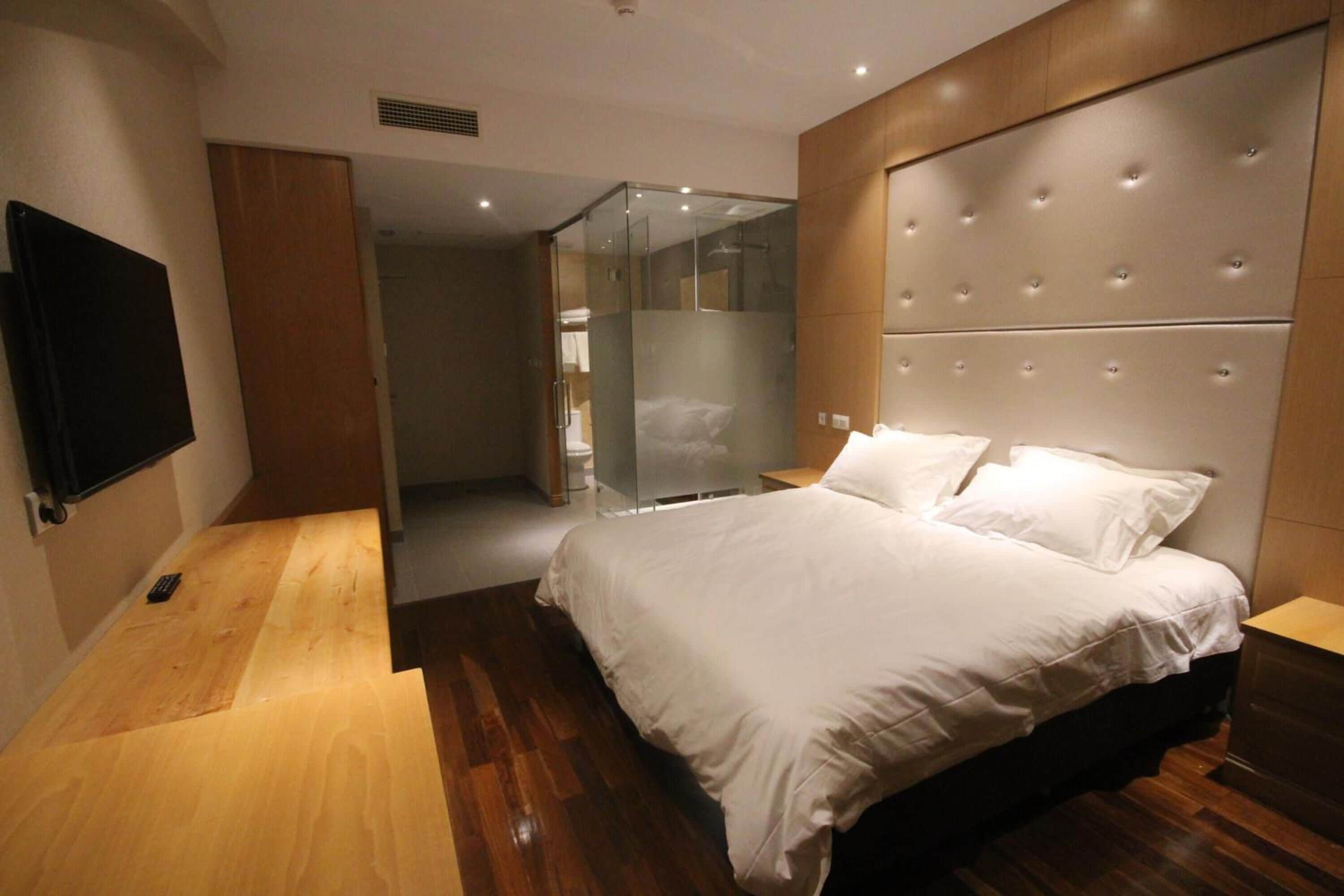 Une chambre d’hôtel moderne avec un plancher en bois, un grand lit avec une literie blanche, une douche en verre et un téléviseur à écran plat fixé au mur.