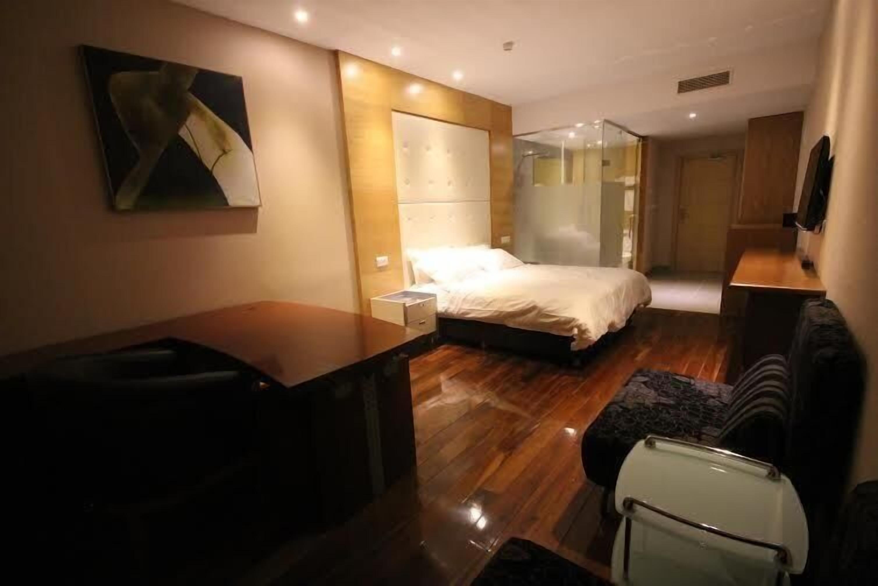 A modern hotel room with a wooden floor, a bed with a white headboard, a small desk, a chair, and a glass shower enclosure.
