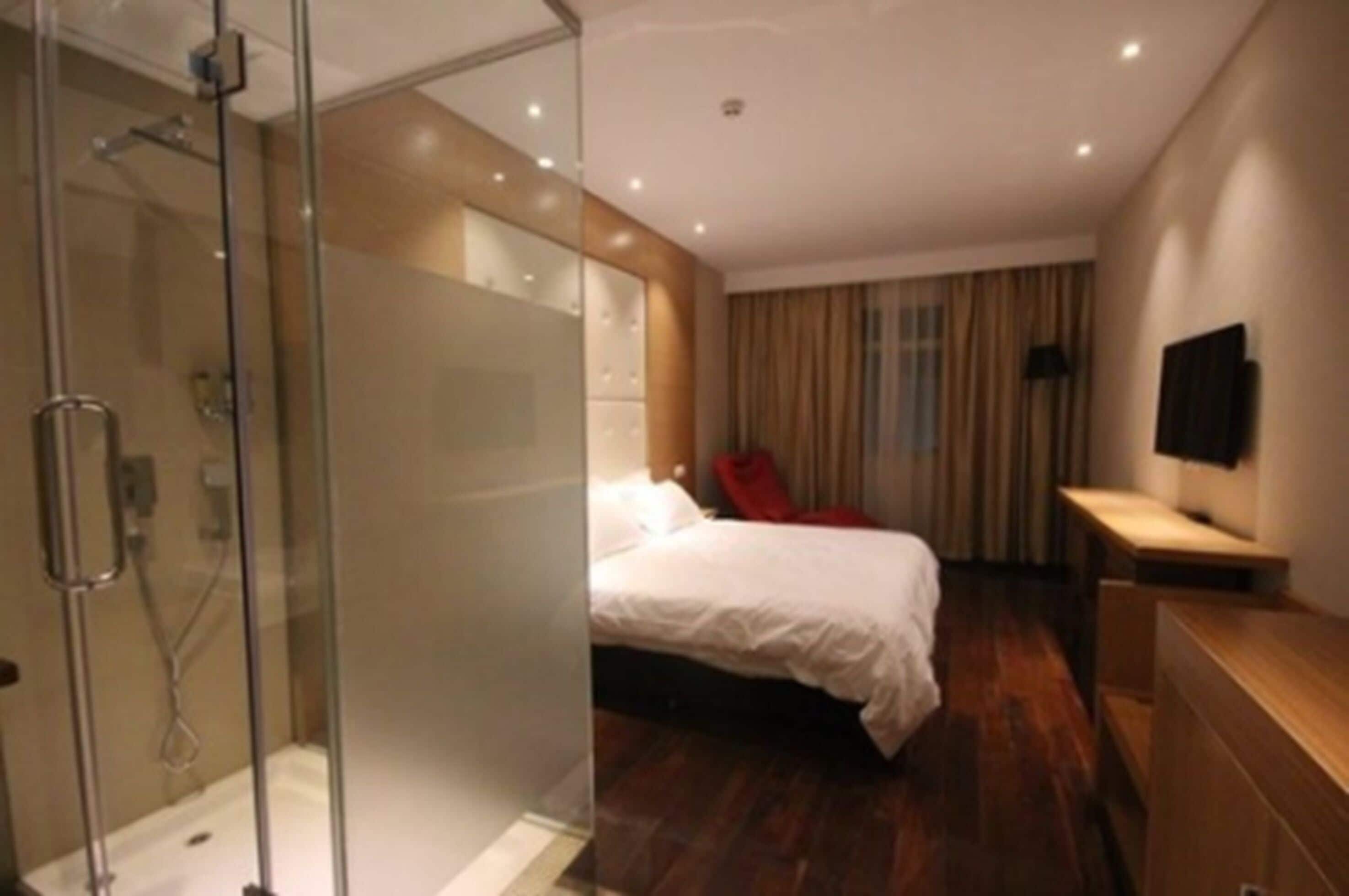 Habitación de hotel moderna con ducha de vidrio, cama con ropa blanca, mesita de noche de madera y televisor de pantalla plana montado en la pared.