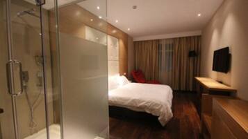 Habitación de hotel moderna con ducha de vidrio, cama con ropa blanca, mesita de noche de madera y televisor de pantalla plana montado en la pared.