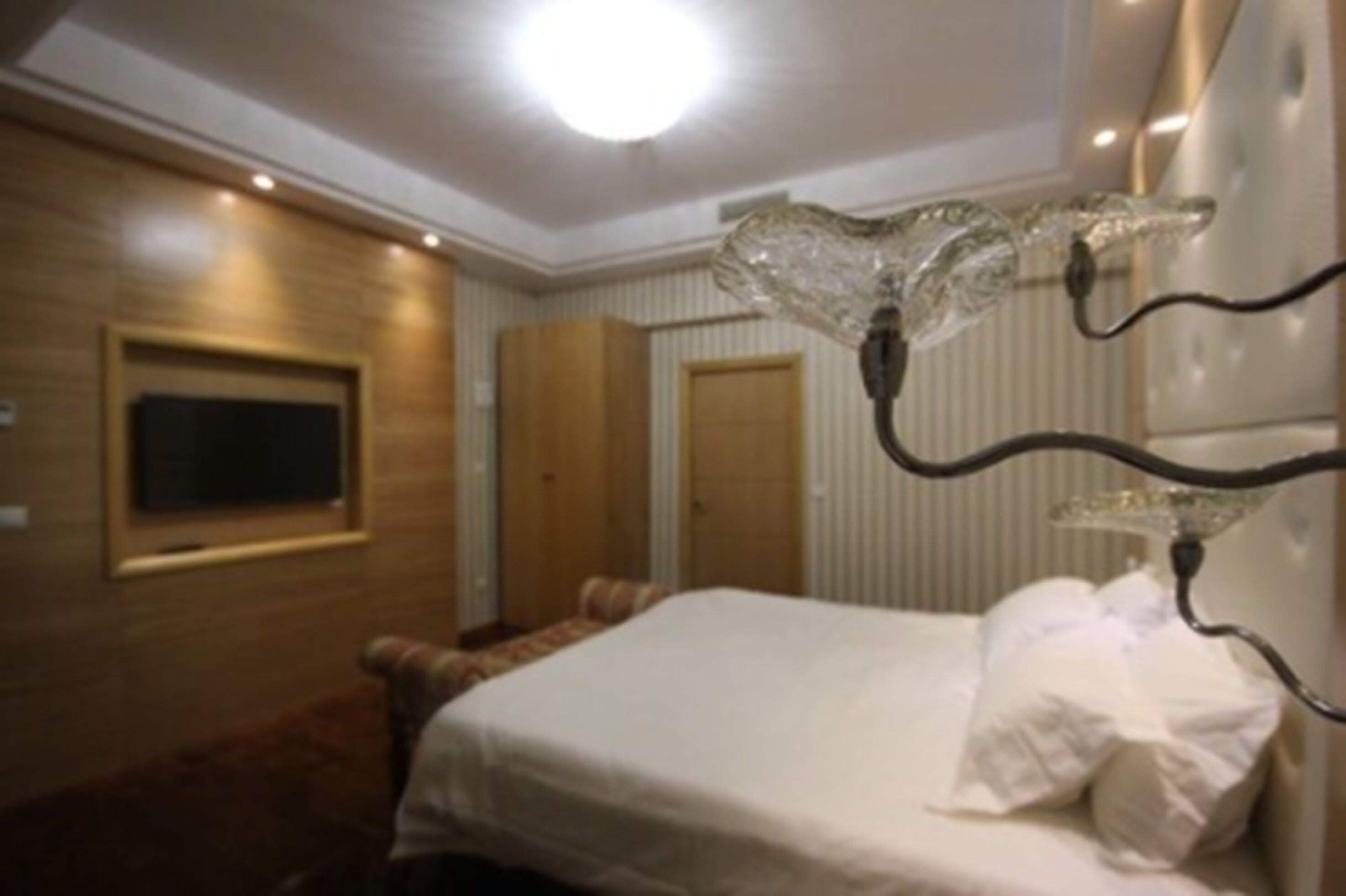 Una habitación de hotel con una cama, un televisor montado en la pared y una lámpara de araña original.