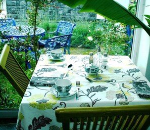 Uma mesa posta para refeição, com toalha de mesa florida, cadeiras azuis e vista de um jardim pela janela.