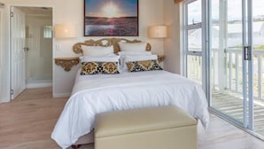 A bedroom with a large bed, two bedside lamps, a wooden headboard, and a view of the beach through a sliding glass door. - Casa Bertella (Yzerfontein)