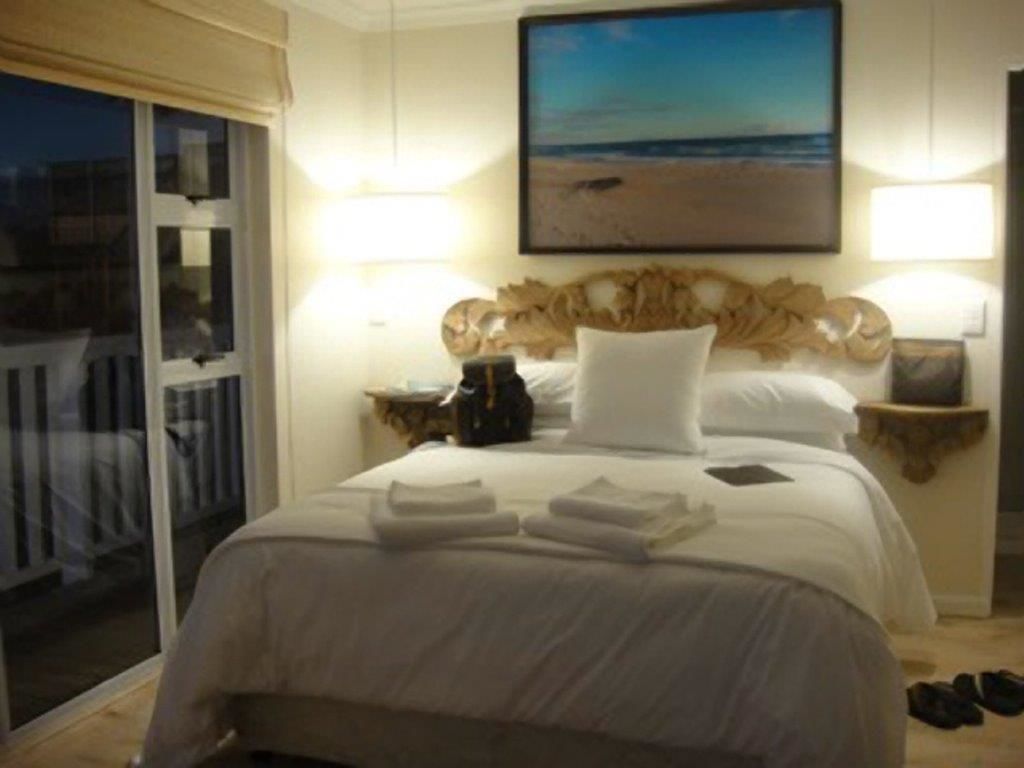 A hotel room with a bed, a nightstand, a lamp, a painting of a beach, and a sliding door to a balcony.