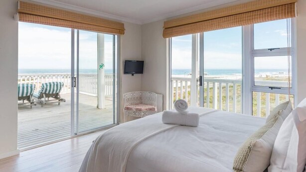A bedroom with a large bed, a chair, and a TV. It has a balcony with a view of the beach. - Casa Bertella (Yzerfontein)