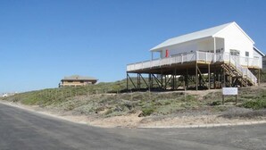 A white building with a balcony on stilts, a clear sky, and a road in the foreground. - Casa Bertella (Yzerfontein)
