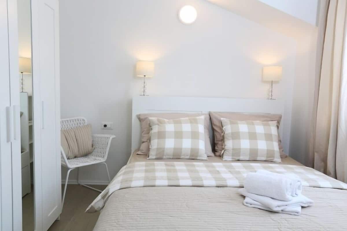 Un dormitorio ordenado con cama, dos lámparas de noche, una silla y una mesita de noche.